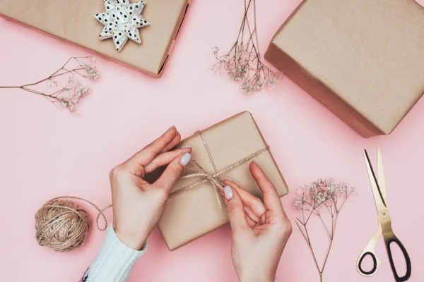 Vista recortada de niña embalaje regalos de Navidad con papel artesanal, cordel y flores, aislado en rosa - foto de stock