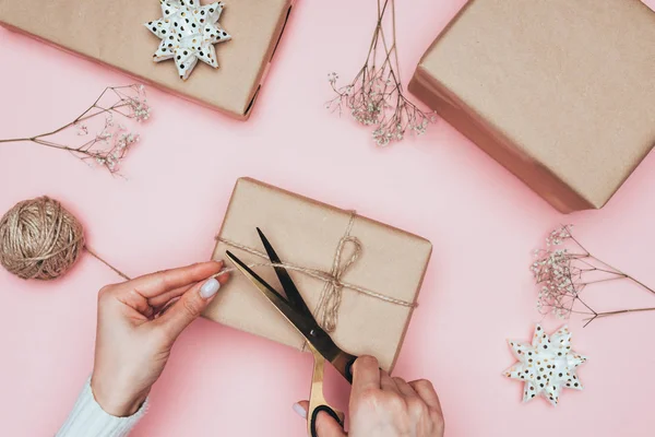 Vista ritagliata di donna con forbici imballaggio regali di Natale con carta artigianale, spago e fiori, isolato su rosa — Foto stock