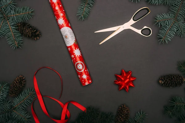 Vista superior del rollo de papel de regalo de Navidad, tijeras, cinta y arco en marco de ramas de abeto y conos de pino en negro - foto de stock