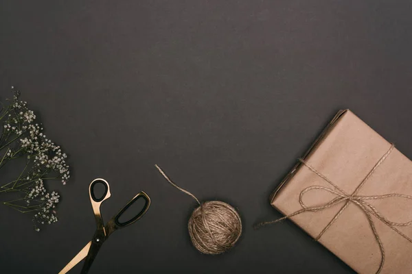 Top view of craft christmas gift with scissors, flowers and twine on black — Stock Photo