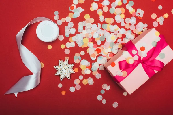 Vue du dessus d'un cadeau de Noël avec arc, ruban et confettis sur rouge — Photo de stock