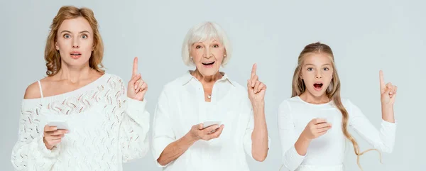 Панорамний знімок шокованої онуки, мати і бабуся тримають смартфони і показують жести ідеї ізольовані на сірому — стокове фото