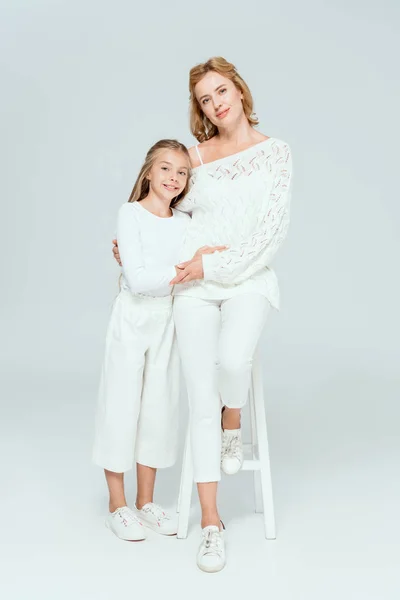 Atractiva madre sentada en el taburete y abrazando sonriente hija sobre fondo gris - foto de stock