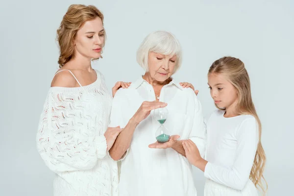 Granddaughter and mother hugging grandmother with hourglass isolated on grey — Stock Photo