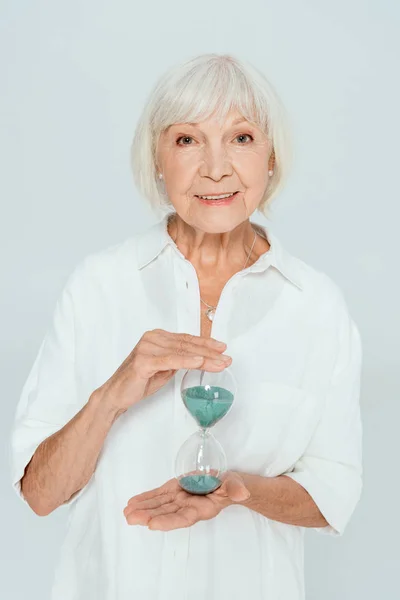 Attractive and smiling woman looking at camera and holding hourglass isolated on grey — Stock Photo