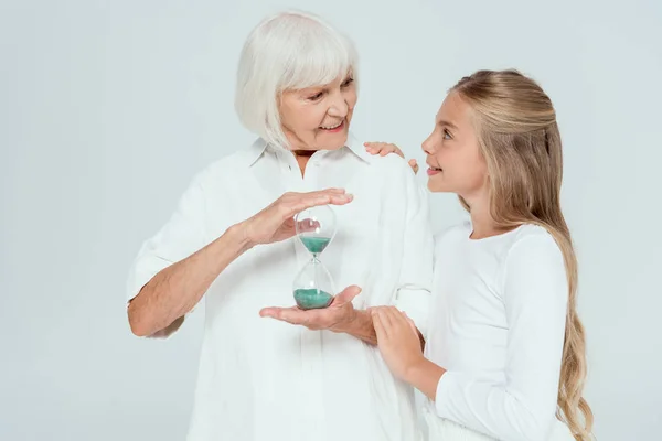 Усміхнена онука обіймає бабусю з пісочним годинником ізольовано на сірому — стокове фото