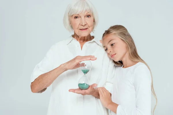 Granddaughter hugging grandmother with hourglass isolated on grey — Stock Photo