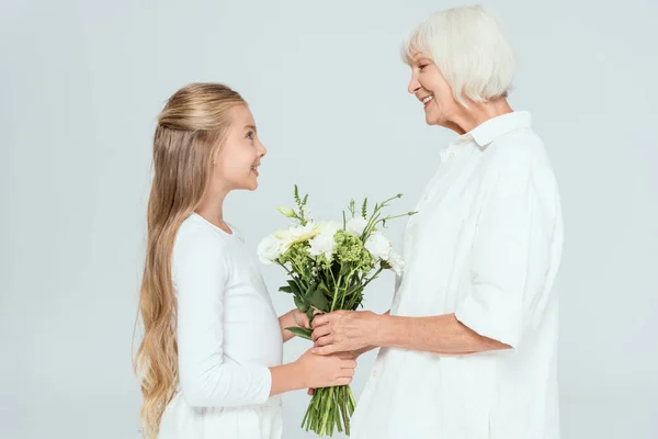 Seitenansicht der lächelnden Enkelin, die Großmutter einen Strauß schenkt, isoliert auf grau — Stockfoto