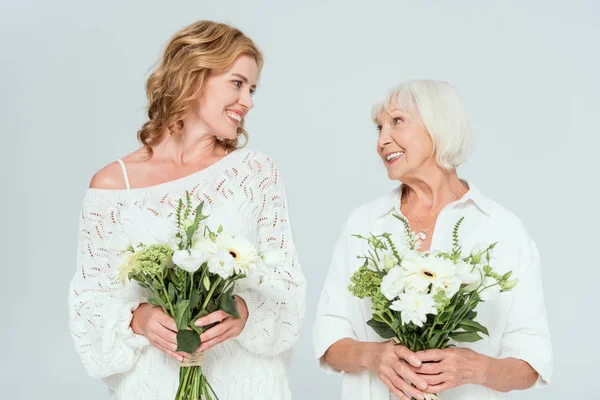 Attraktive Mutter und lächelnde Tochter halten Blumensträuße in der Hand und betrachten einander isoliert auf grau — Stockfoto