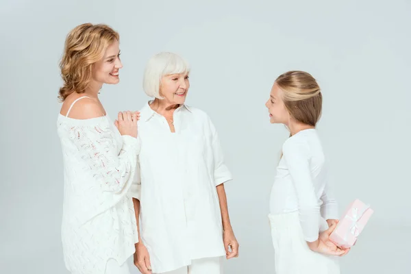 Souriant petite-fille tenant cadeau et regardant mère et grand-mère isolées sur gris — Photo de stock