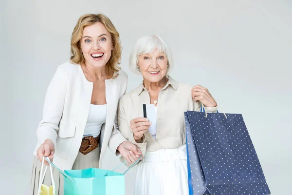 Sonriente madre con tarjeta de crédito e hija sosteniendo bolsas aisladas en gris - foto de stock