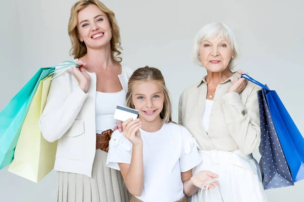 Улыбающаяся внучка с кредиткой, бабушка и мама с сумками для покупок, изолированными на сером — стоковое фото