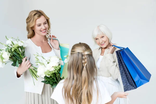 Back view of granddaughter holding bouquet and looking at grandmother and mother with shopping bags isolated on grey — Stock Photo