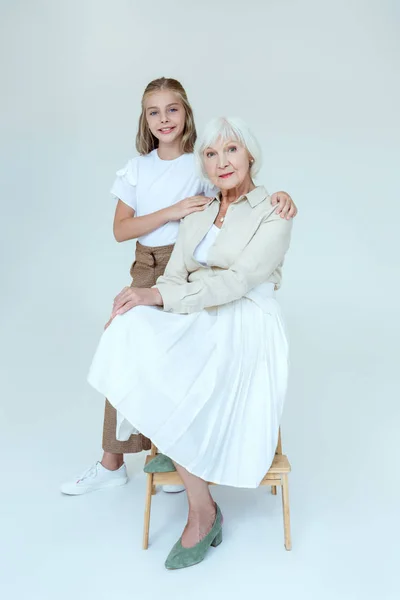Улыбающаяся внучка обнимает бабушку на стуле на сером фоне — стоковое фото