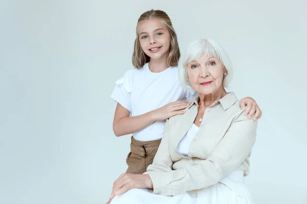 Sonriente y linda nieta abrazando abuela aislado en gris - foto de stock