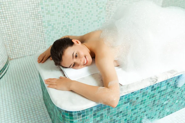 Vista de ángulo alto de la mujer sonriente acostada en la mesa de hammam con espuma en baño turco - foto de stock