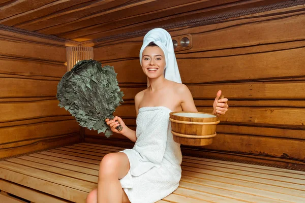 Donna sorridente in asciugamani in possesso di scopa di betulla e vasca da bagno in sauna — Foto stock