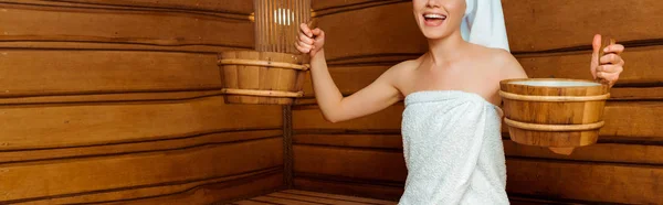 Panoramic shot of smiling woman in towels holding washtubs in sauna — Stock Photo