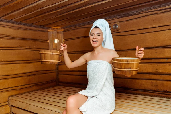 Mujer sonriente y atractiva en toallas sosteniendo bañeras en sauna - foto de stock