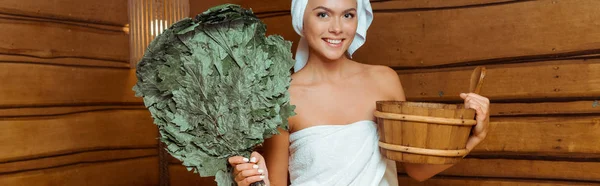 Panoramic shot of smiling and attractive woman in towels holding washtub and birch broom in sauna — Stock Photo