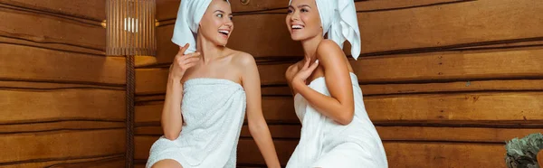 Panoramic shot of smiling and attractive friends in towels in sauna — Stock Photo