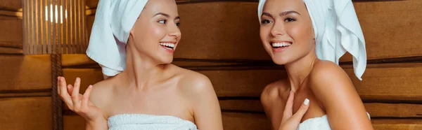 Plano panorámico de amigos sonrientes y atractivos en toallas hablando en sauna - foto de stock