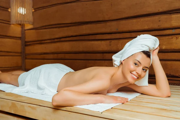 Привлекательная и улыбающаяся женщина в полотенцах, лежащих в сауне — стоковое фото