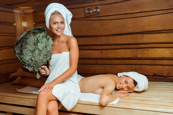Femme souriante et attrayante tenant balai de bouleau et son ami couché dans le sauna — Photo de stock