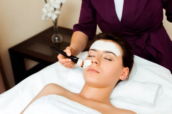Abgeschnittene Ansicht von Kosmetikerin, die Gesichtsmaske auf attraktive Frau im Spa aufträgt — Stockfoto