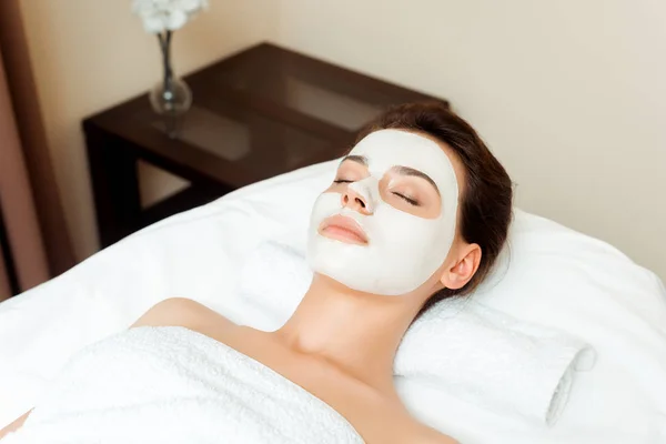 Vista de ángulo alto de la mujer atractiva con máscara en la cara acostado en la mesa de masaje en el spa - foto de stock