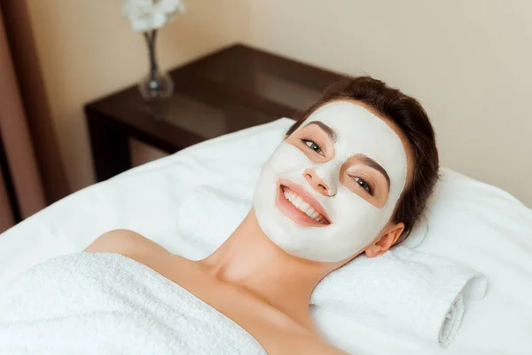 Vue grand angle de la femme souriante avec masque sur le visage couché sur la table de massage dans le spa — Photo de stock