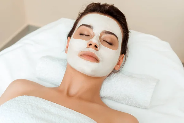 Vista ad alto angolo di donna attraente con maschera sul viso sdraiato sul tavolo da massaggio in spa — Foto stock