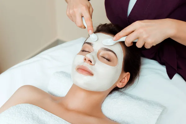 Vista recortada del cosmetólogo aplicando mascarilla facial en mujer atractiva en spa - foto de stock