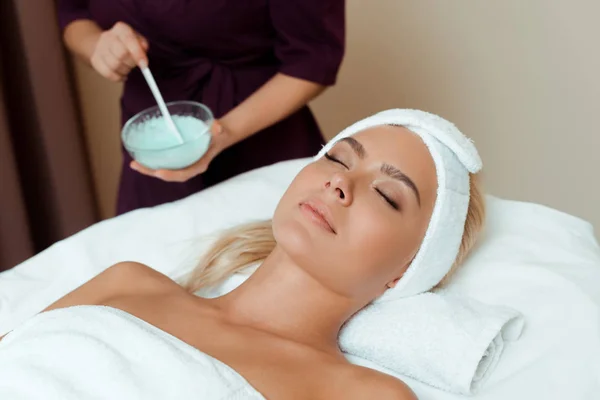 Vista ritagliata di cosmetologo in possesso di ciotola con maschera viso e attraente donna sdraiata in spa — Foto stock