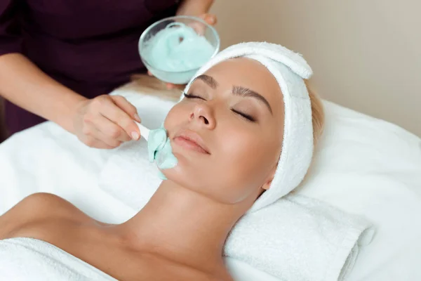 Vista recortada del cosmetólogo aplicando mascarilla facial en mujer atractiva en spa - foto de stock