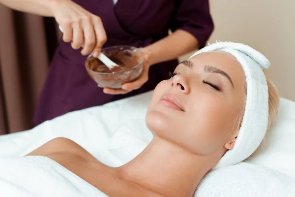 Vista ritagliata di cosmetologo in possesso di ciotola con maschera viso e attraente donna sdraiata in spa — Foto stock