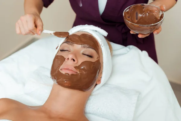 Vista recortada del cosmetólogo aplicando mascarilla facial en mujer atractiva en spa - foto de stock