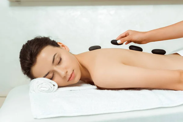 Vista cortada de massagista fazendo massagem de pedra quente para mulher atraente no spa — Fotografia de Stock