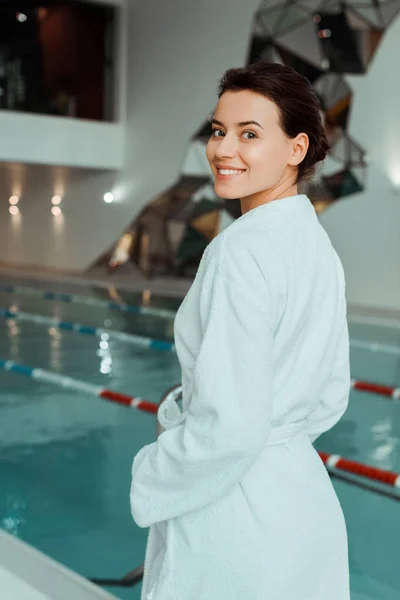 Attraktive und lächelnde Frau im weißen Bademantel, die im Wellnessbereich in die Kamera schaut — Stockfoto
