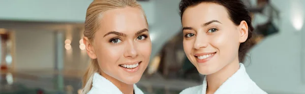 Panoramic shot of attractive and smiling friends in white bathrobes  looking at camera in spa — Stock Photo