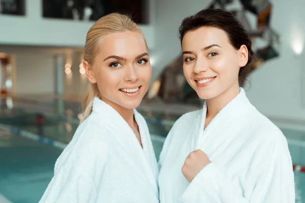 Attractive and smiling friends in white bathrobes looking at camera in spa — Stock Photo