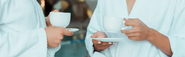 Panoramic shot of friends in white bathrobes holding cups in spa — Stock Photo