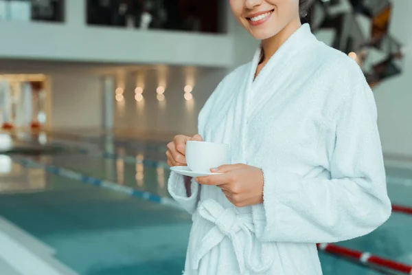 Обрізаний вид усміхненої жінки в білому халаті, що тримає чашку кави в спа — стокове фото