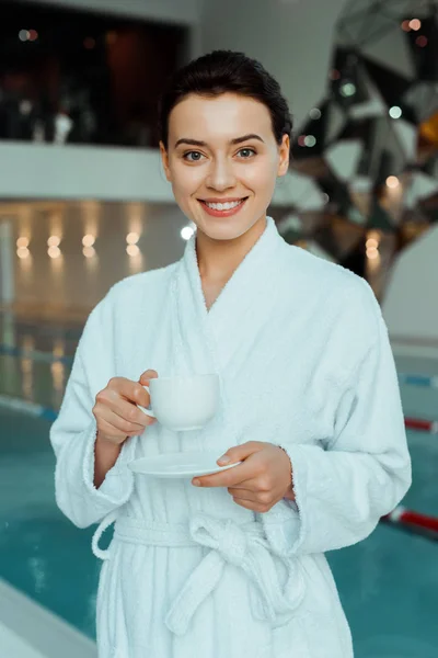 Mulher atraente e sorridente em roupão de banho branco segurando xícara de café no spa — Fotografia de Stock