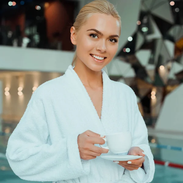 Привлекательная и улыбающаяся женщина в белом халате с чашкой кофе в спа — стоковое фото