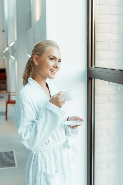Attractive and smiling woman in white bathrobe holding cup of coffee and looking through window in spa — Stock Photo
