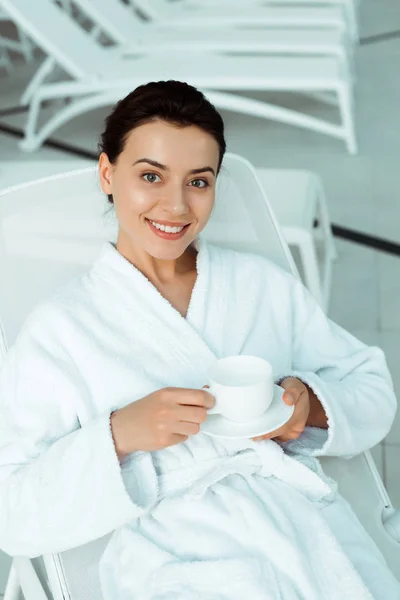 Vista ad alto angolo di donna attraente e sorridente in accappatoio bianco con una tazza di caffè in spa — Foto stock