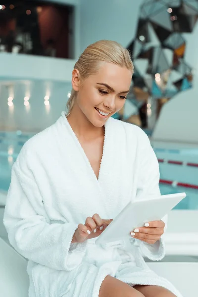 Femme attrayante et souriante en peignoir blanc en utilisant une tablette numérique dans le spa — Photo de stock