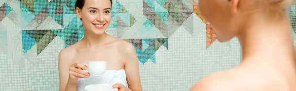 Панорамный снимок привлекательной и улыбающейся женщины в белом полотенце, разговаривающей с другом и держащей чашку в спа — стоковое фото