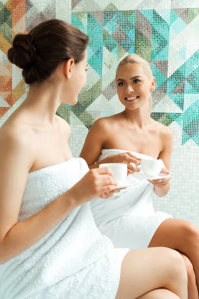 Amigos atractivos y sonrientes en toallas blancas hablando y sosteniendo tazas en el spa - foto de stock